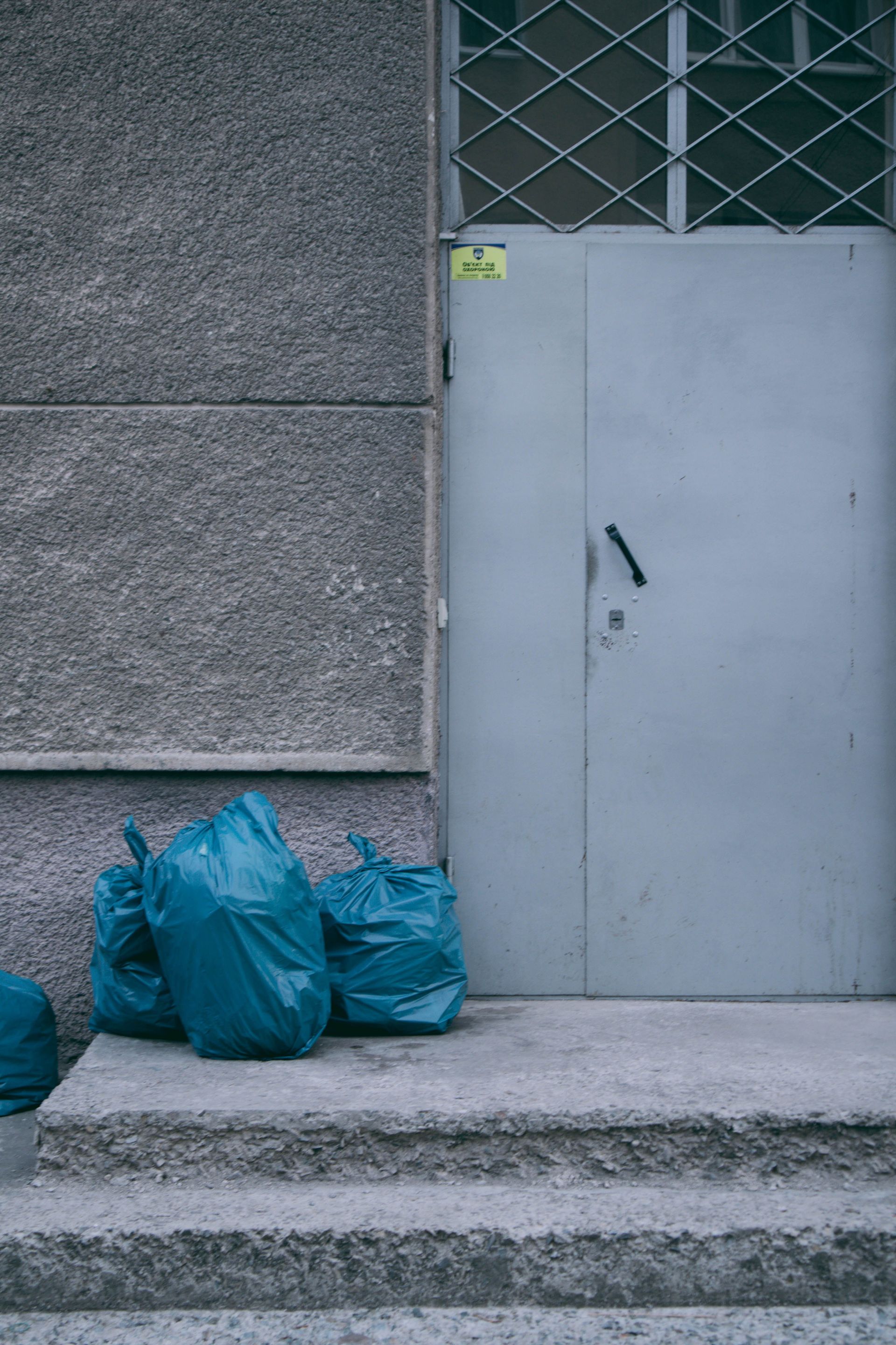 A quiet urban scene featuring blue garbage bags by a metal door on concrete steps.